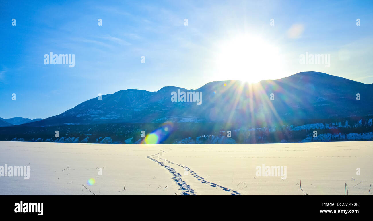 Stampe di piedi nella neve su un lago ghiacciato con sun flare e montagne coperte di neve in background. Un inverno paesaggio di montagna in British colonna Foto Stock