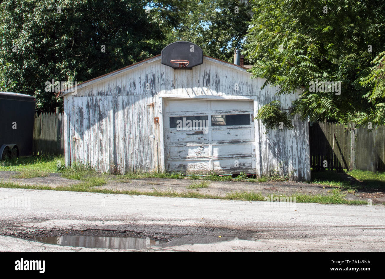 Vecchio garage in legno si siede dimenticato in una pentola forata di street, con un antico Basketball hoop appeso sopra la vernice della porta di pelatura Foto Stock
