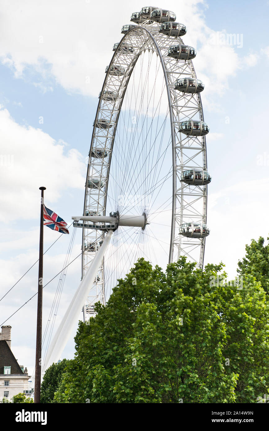 Vista del London Eye con la bandiera britannica e spiegata verdi alberi in primo piano. Bella giornata di sole e soffici nuvole. Copia dello spazio. Foto Stock