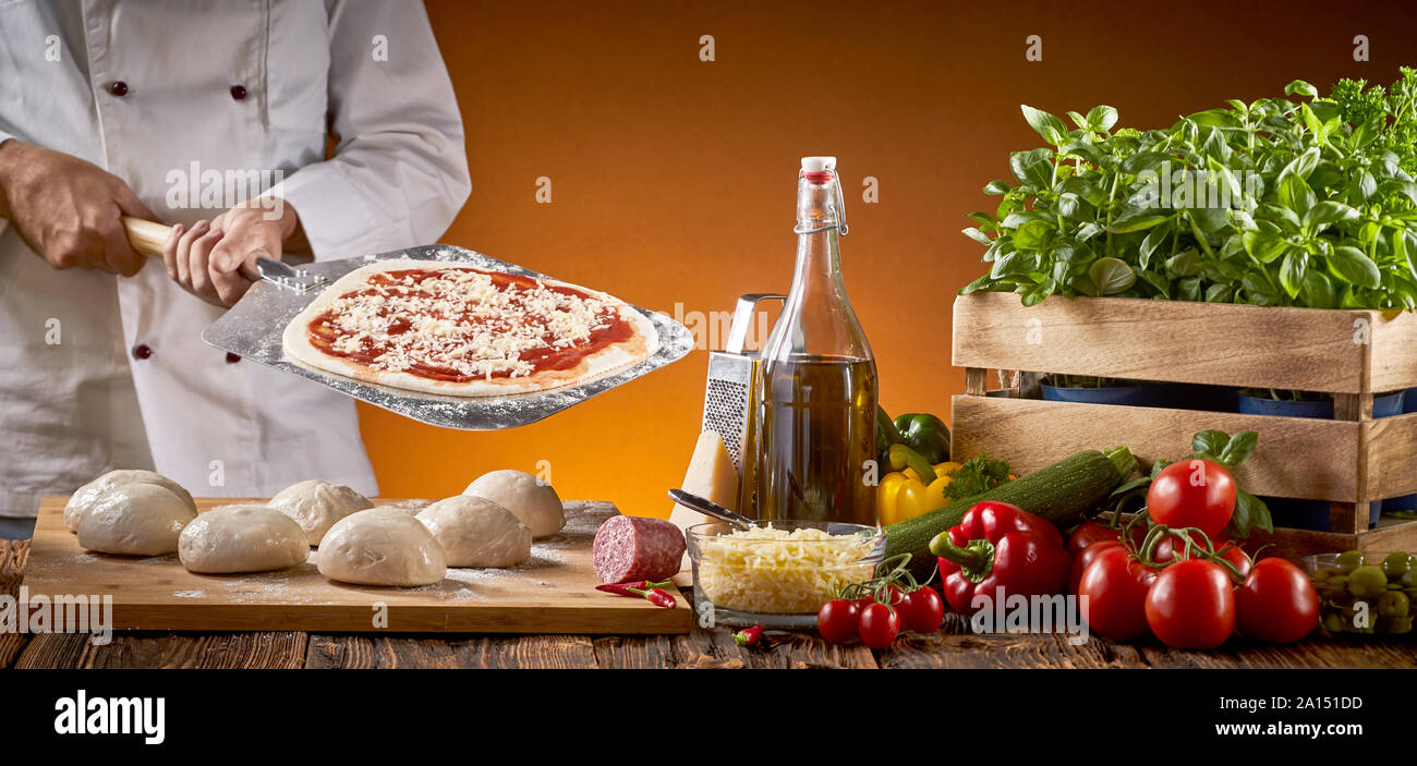 Cuoco preparare una pizza italiana in un ristorante in possesso di un dispositivo di compressione con base di pasticceria dietro un tavolo rustico con assortimento di ingredienti freschi e di porzioni o Foto Stock