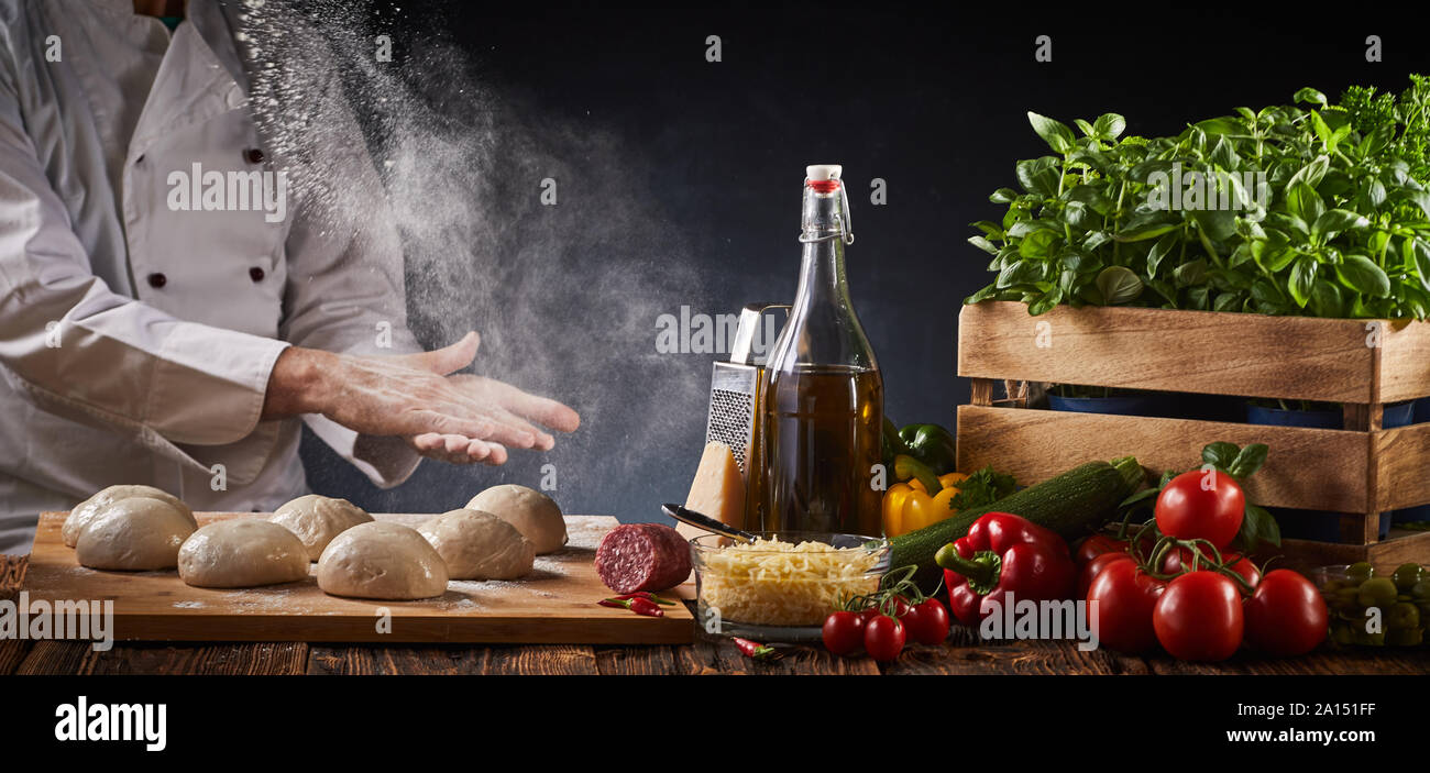 Lo chef prepara una specialità fatte in casa pizza italiana battendo le mani in una nuvola di farina al di sopra di porzioni di impasto crudo su una tabella con fresca assortita ho Foto Stock