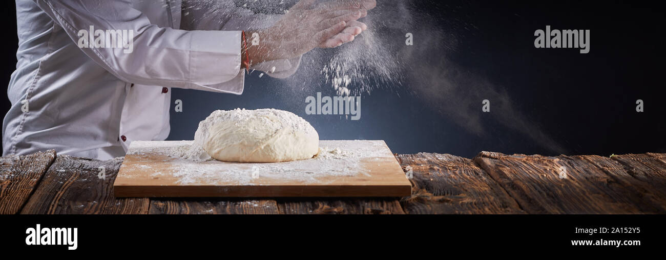 Banner di panorama di uno chef battendo le mani con una nuvola di farina in aria come polveri di lui un tumulo di pasta cruda su un bordo mentre facendo pizze o Foto Stock