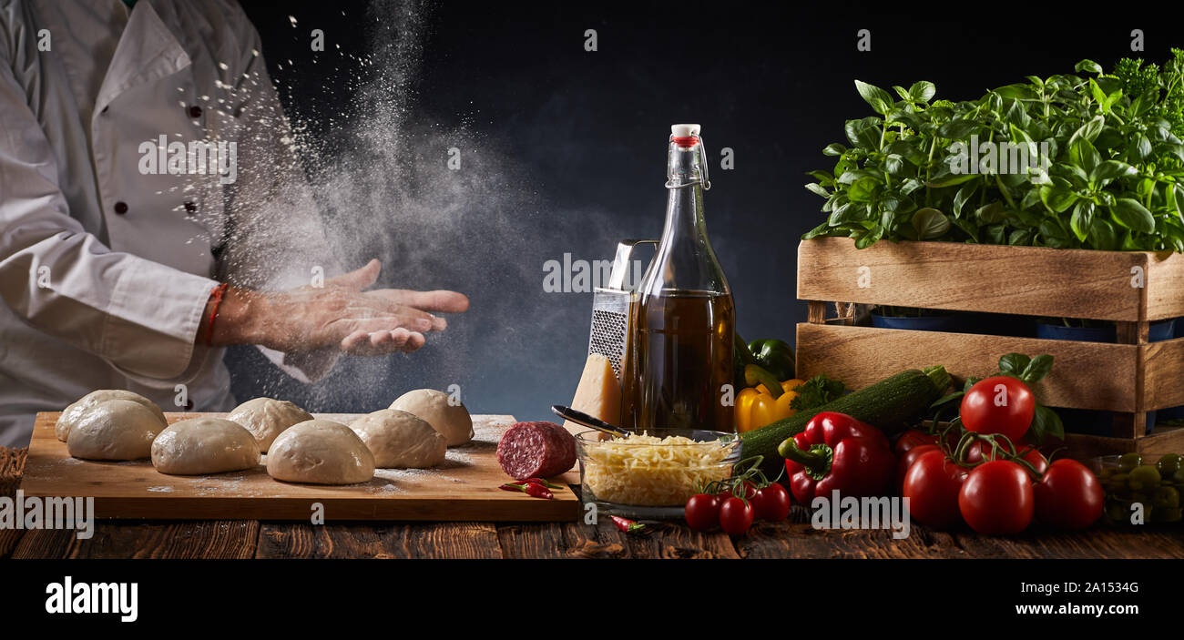 Lo Chef battendo le mani con la farina sospesa in un blocco motion view come egli pone di porzioni di impasto crudo al fianco di freschi ingredienti della pizza su un rustico Foto Stock
