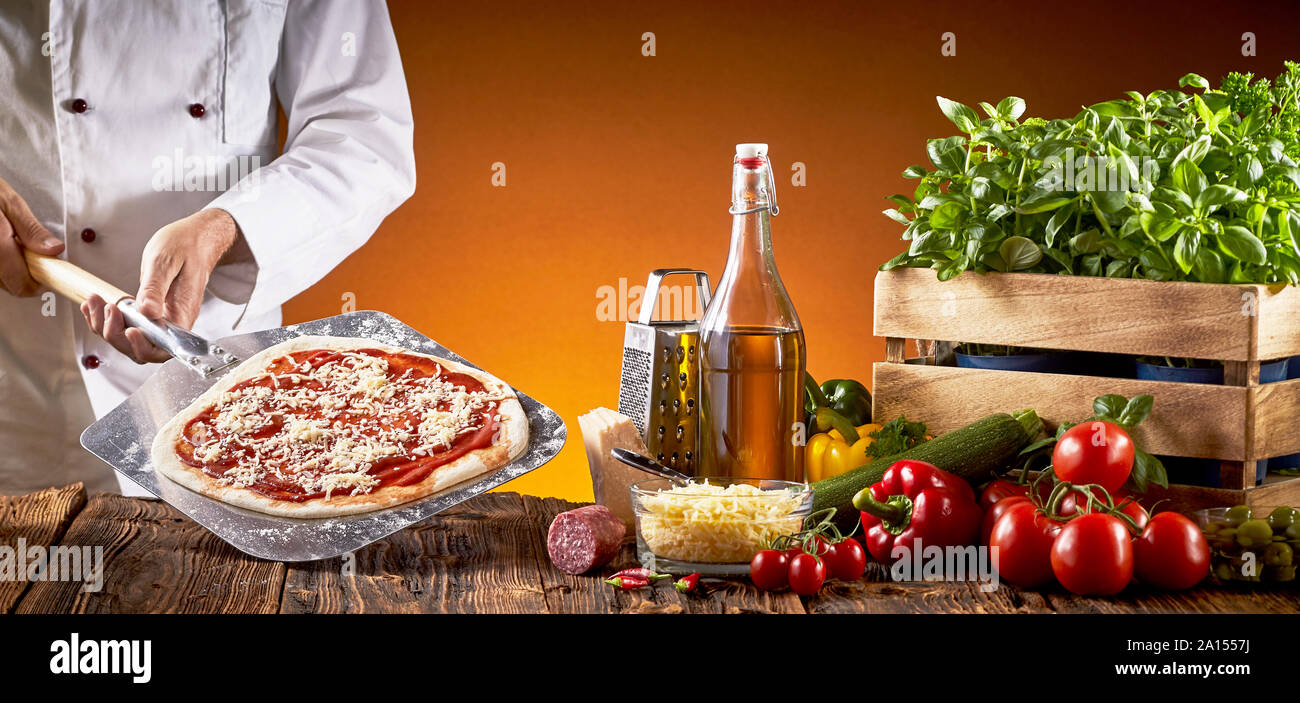 Lo Chef facendo una tradizionale pizza italiana in una pizzeria tenendo una torta base con pomodoro e formaggio su una paletta con assortimento di ingredienti freschi per un bro Foto Stock