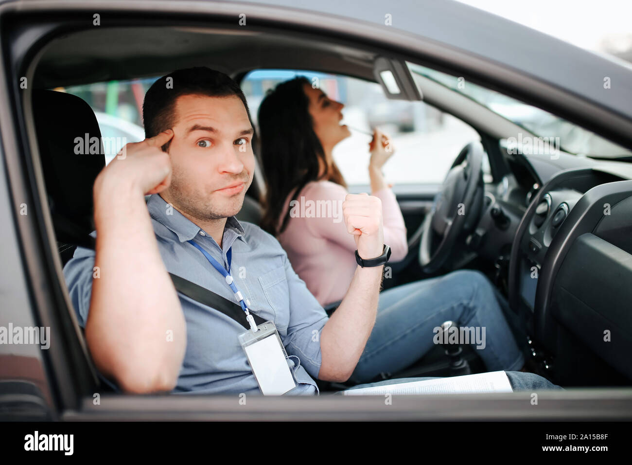 Maschio istruttore auto prende in esame giovane donna. Cercare sulla fotocamera e punto sulla testa con il dito. Mad studente facendo trucco in auto. Guardare sullo specchio Foto Stock