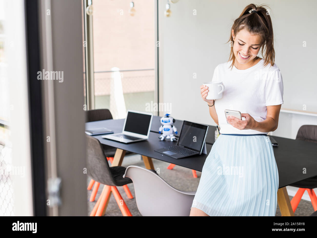 Sorridente giovane imprenditrice avente pausa caffè in ufficio guardando al telefono cellulare Foto Stock