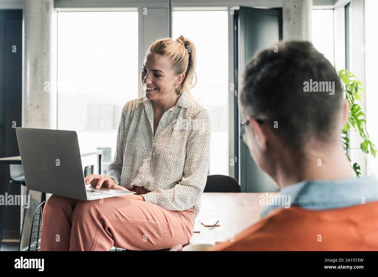 Sorridente imprenditrice seduta sul tavolo in ufficio utilizzando laptop con imprenditore in primo piano Foto Stock