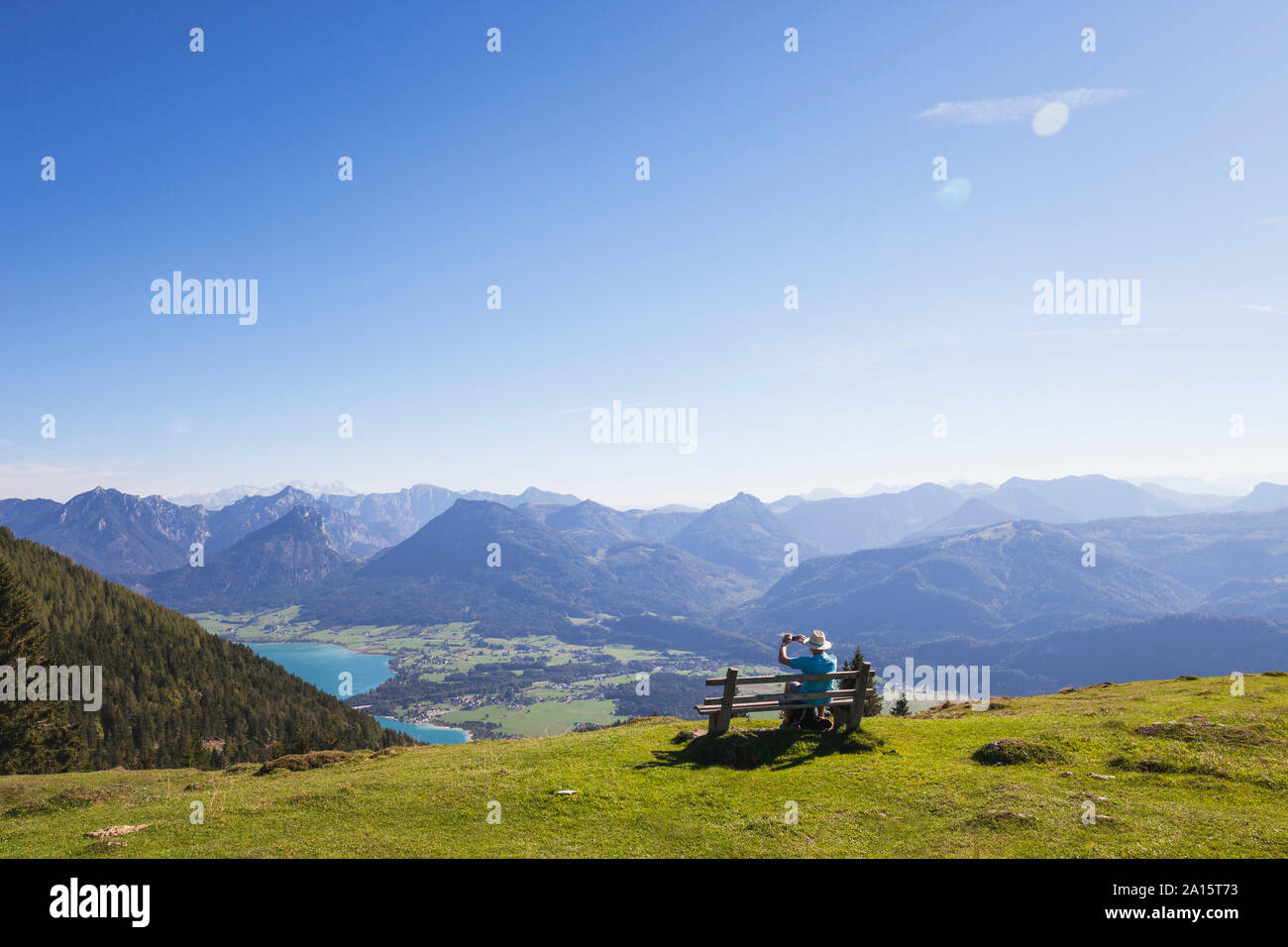 Vista posteriore del maschio senior escursionista fotografare mentre è seduto su un banco mentre guardando Dachstein montagne contro il cielo blu Foto Stock