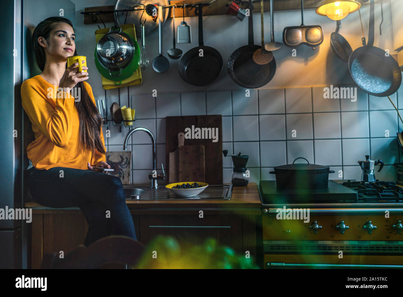 Giovane donna seduta sul banco di cucina a casa gustando un caffé Foto Stock