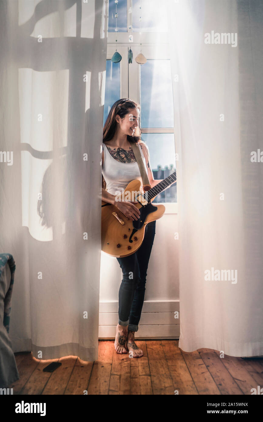 Felice giovane donna in piedi presso la finestra a casa con una chitarra Foto Stock