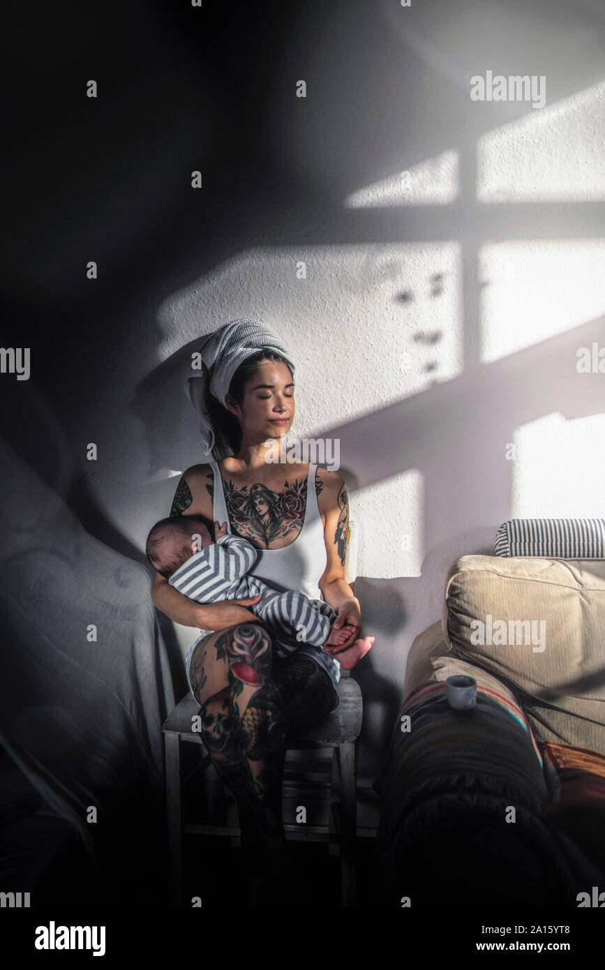 Tatuato giovane donna tenendo il suo bambino a casa godendo il sole Foto Stock