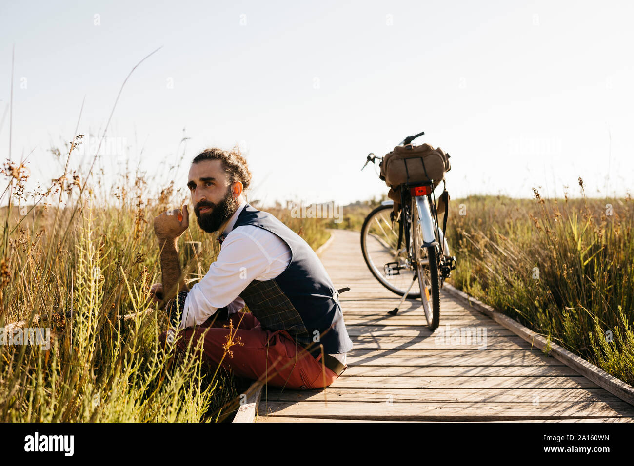 Ben vestito uomo seduto su una passerella di legno in campagna accanto a una bicicletta Foto Stock