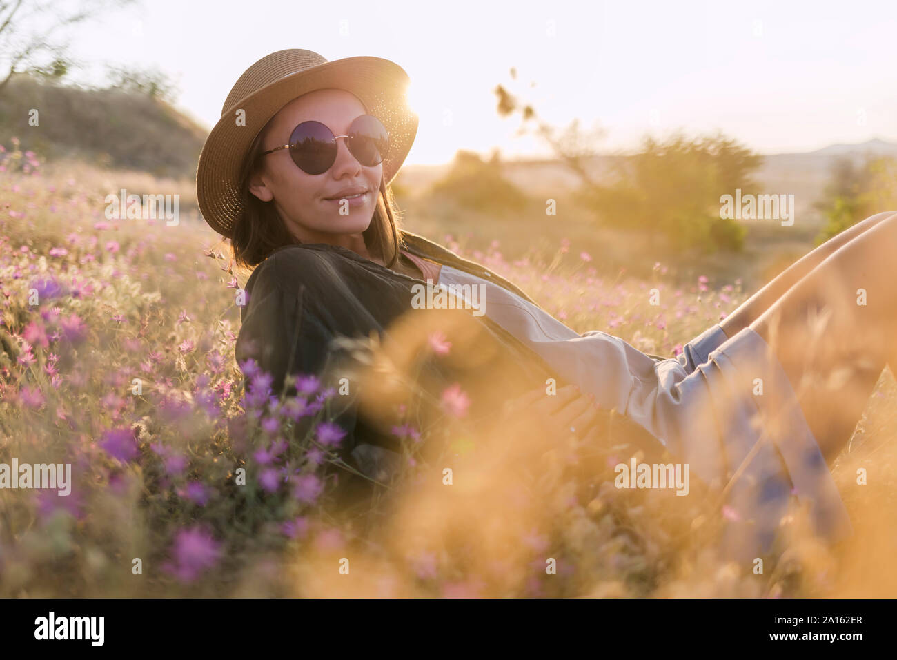 Giovane donna che indossa gli occhiali da sole e cappello, seduto sul prato al tramonto Foto Stock