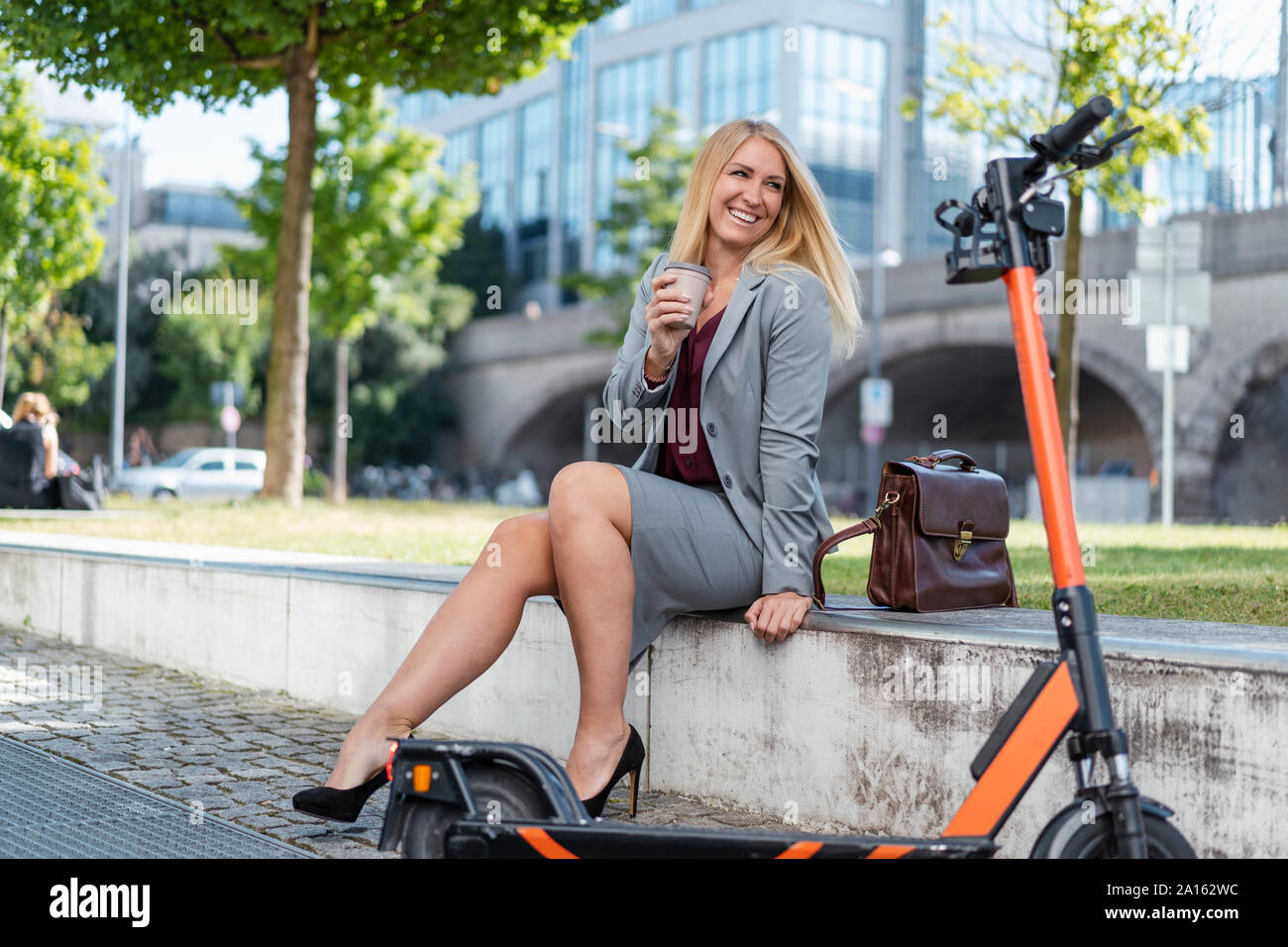 Ritratto di bionda imprenditrice con lo scooter elettrico seduto su una parete di bere il caffè per andare Foto Stock