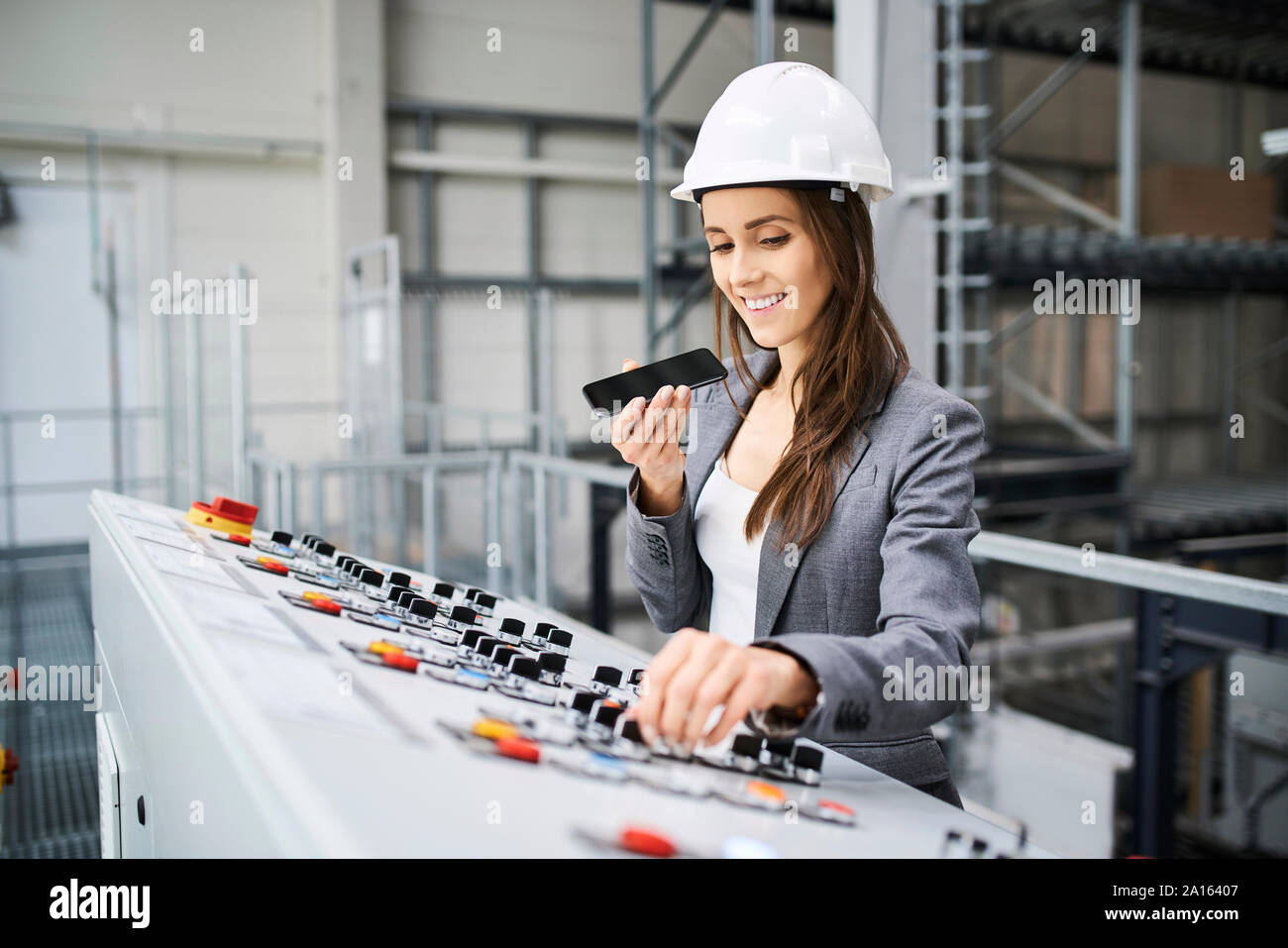 Donna che indossa elmetto in pannello di controllo in una fabbrica tramite telefono cellulare Foto Stock