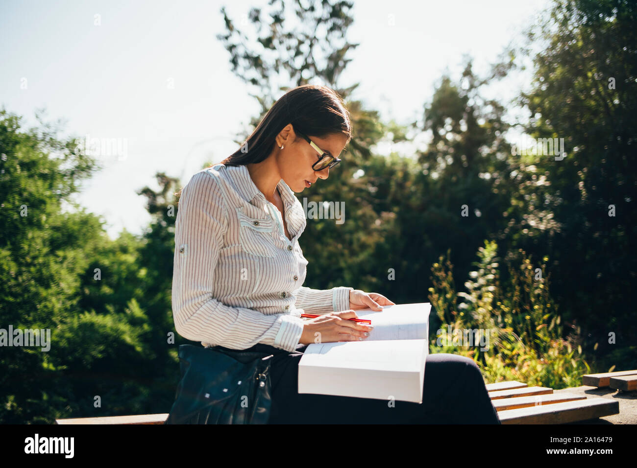 Giovane donna seduta su una panchina in un parco guardando un libro aperto Foto Stock