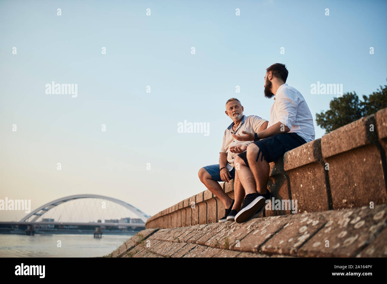Padre e figlio adulto seduto su una parete in riva al fiume e a parlare Foto Stock