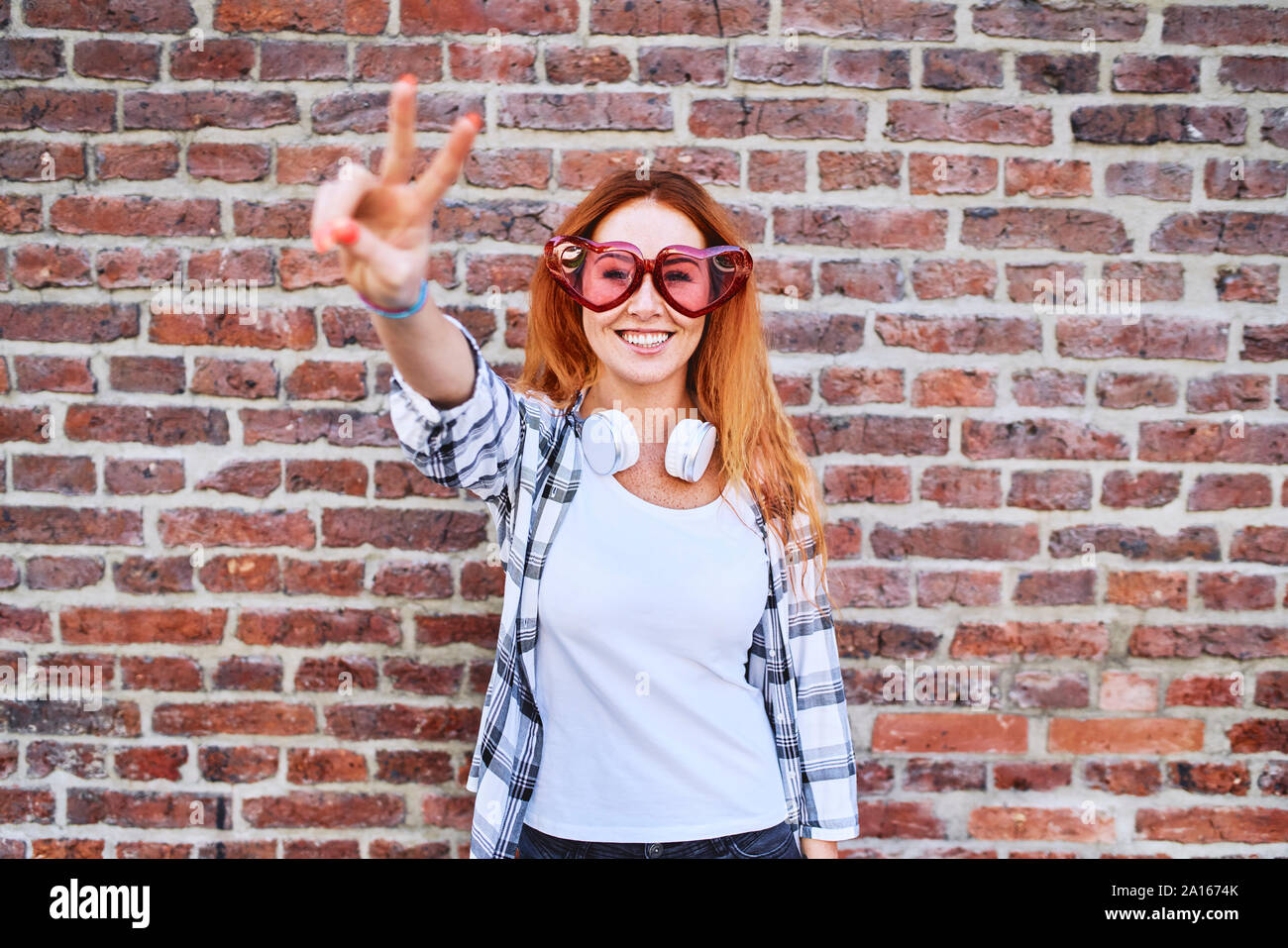 Stupendo giovane donna in forma di cuore bicchieri sorridente alla fotocamera e compiendo il gesto di pace Foto Stock