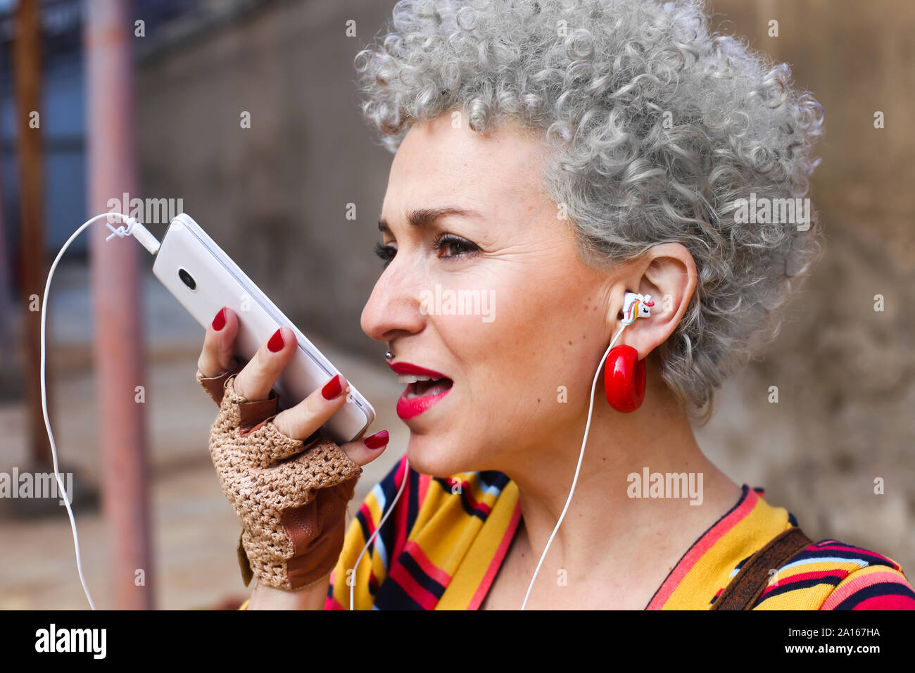 Ritratto di trafitto donna matura con il grigio capelli ricci utilizzando auricolari e il telefono cellulare Foto Stock