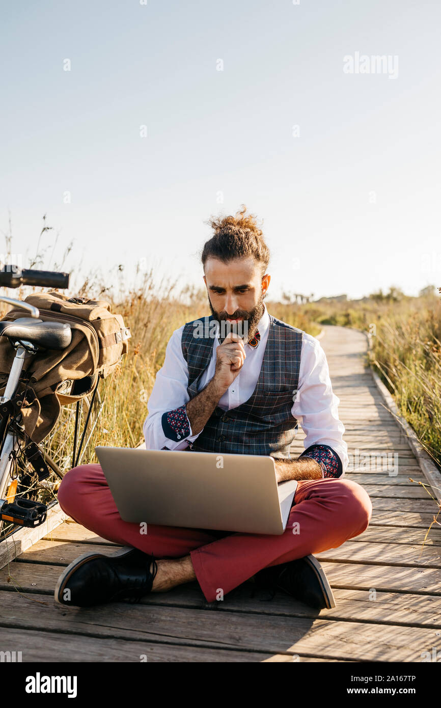 Ben vestito uomo seduto su una passerella di legno in campagna accanto a una bicicletta utilizzando laptop Foto Stock