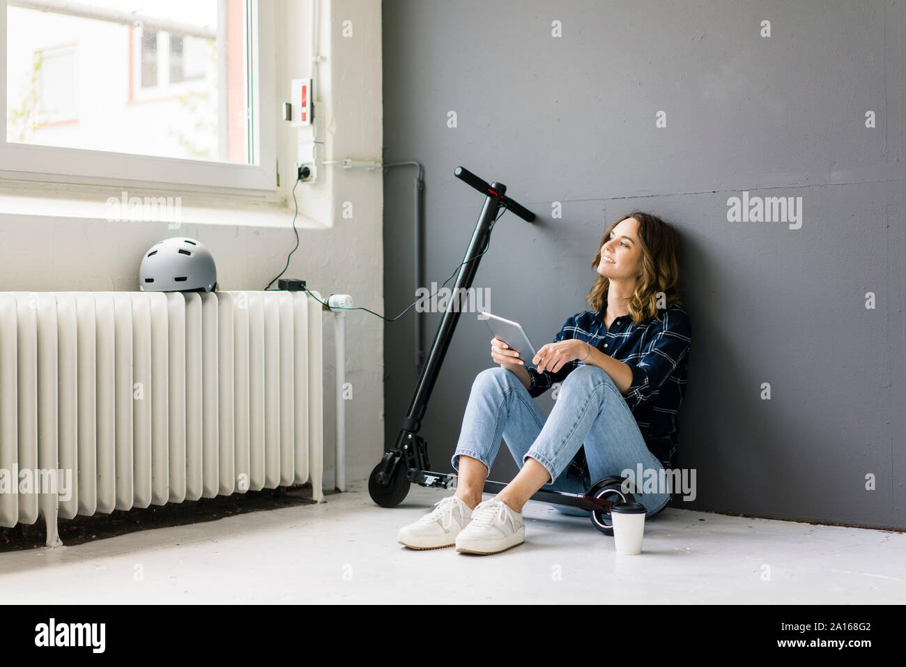 Giovane donna con e-scooter seduto sul pavimento, con tavoletta digitale Foto Stock
