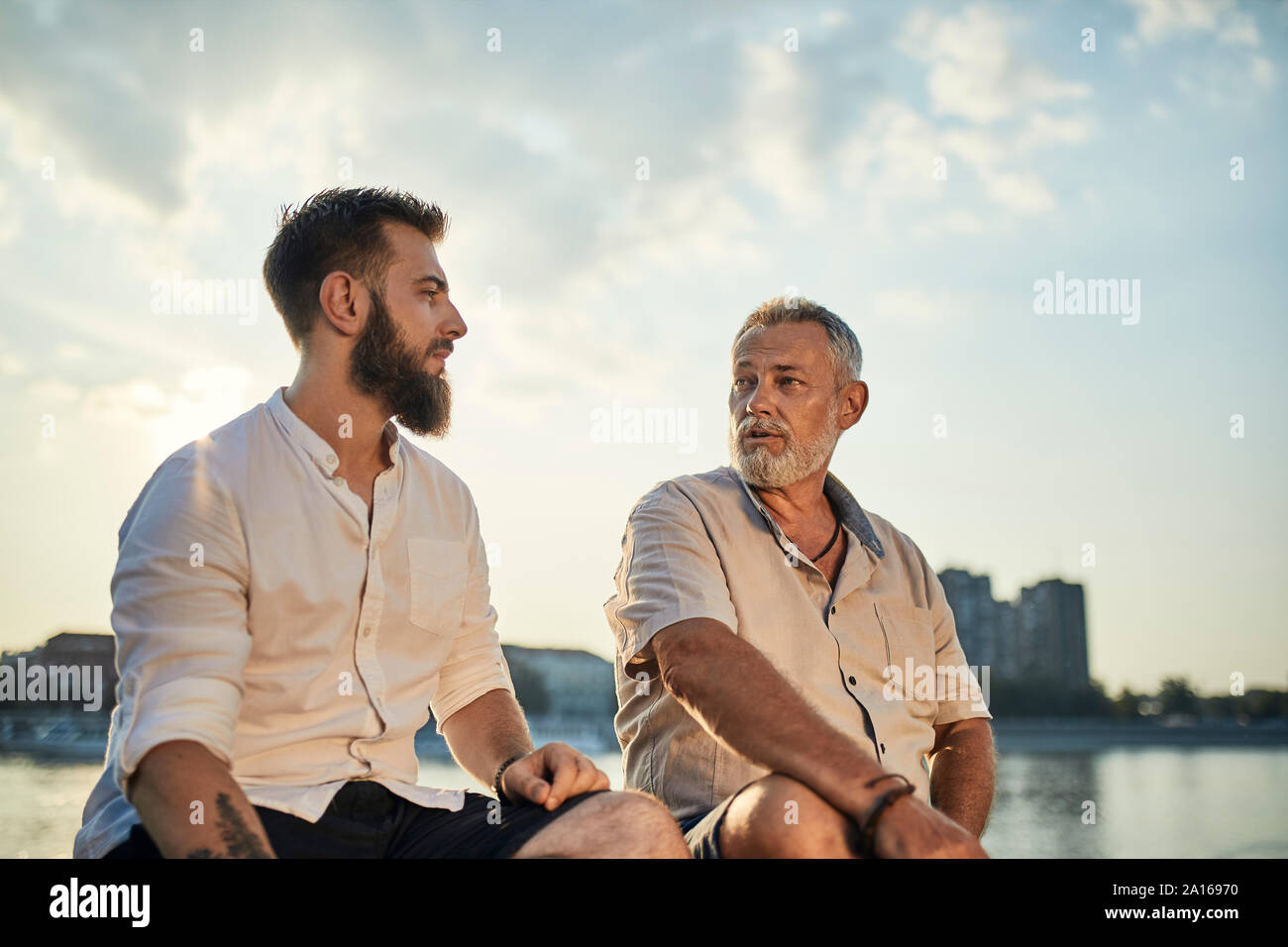 Padre e figlio adulto seduto su una parete in riva al fiume e a parlare Foto Stock