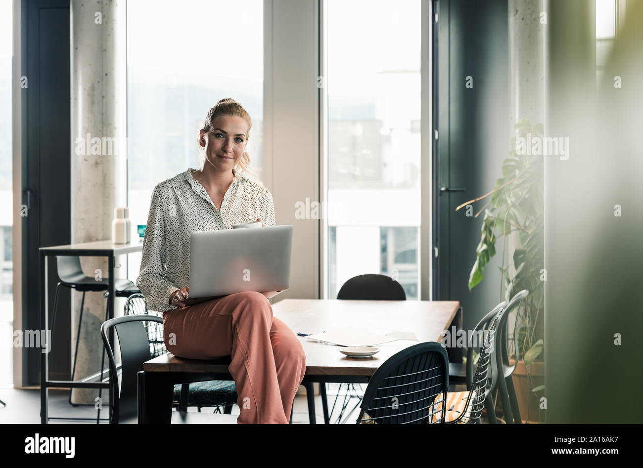 Ritratto di imprenditrice seduta sul tavolo in ufficio con computer portatile Foto Stock