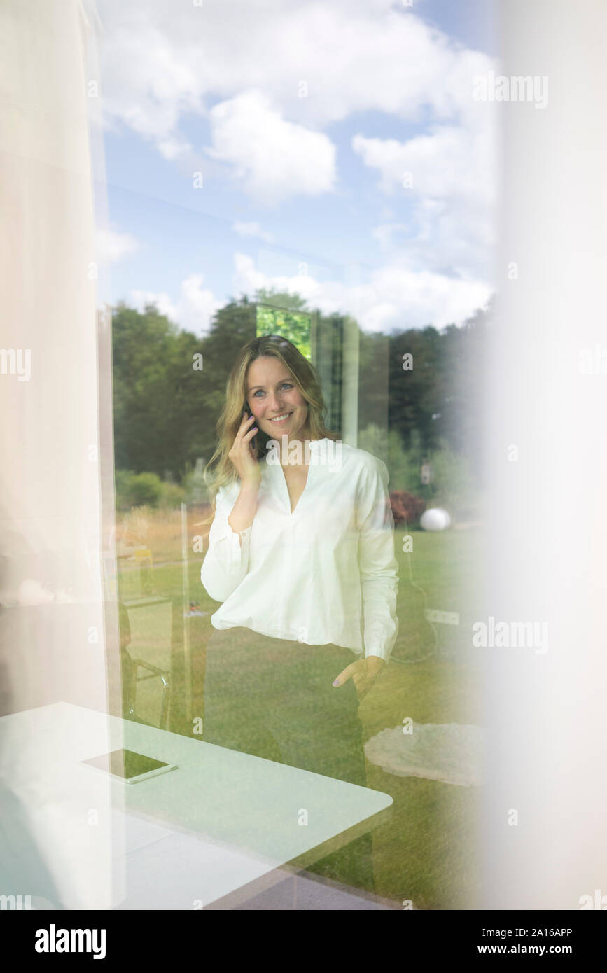 Sorridente imprenditrice parlando al telefono a casa dietro il vetro di finestra Foto Stock