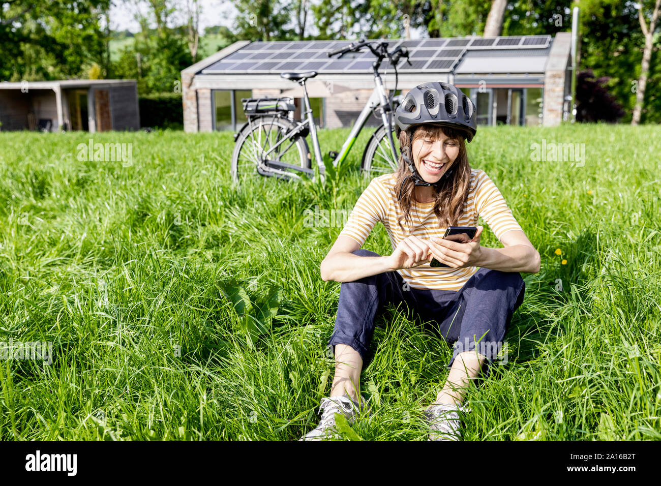 Ridendo donna con bicicletta utilizzando il telefono cellulare su un prato di fronte a casa Foto Stock