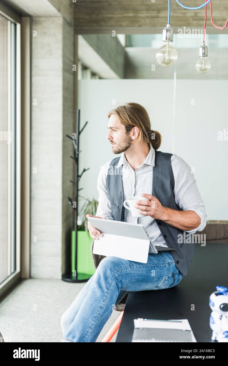 Giovane imprenditore seduta sul tavolo in ufficio con tavoletta e la tazza di caffè Foto Stock
