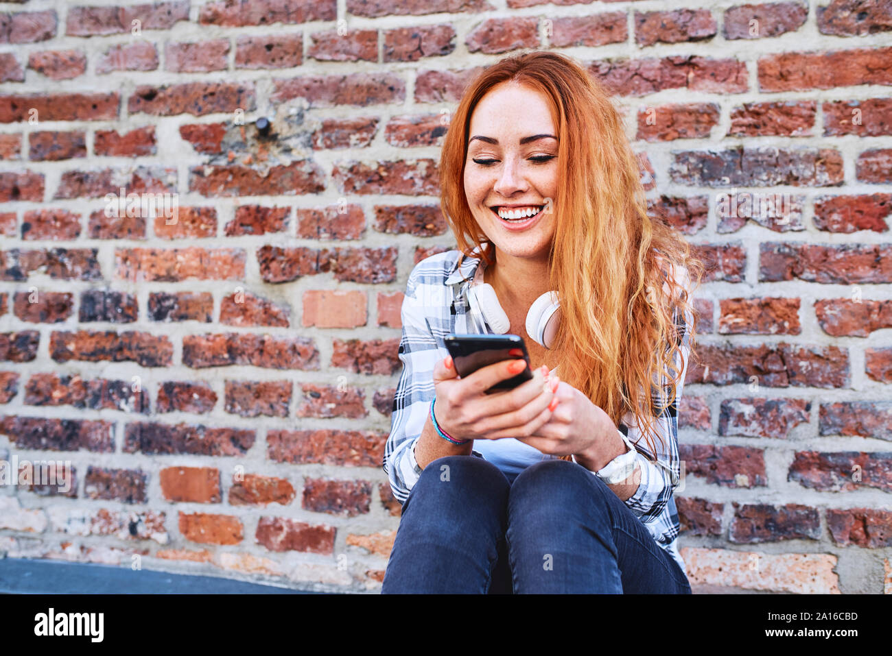 Allegro giovane donna seduta contro un muro di mattoni e utilizza lo smartphone Foto Stock