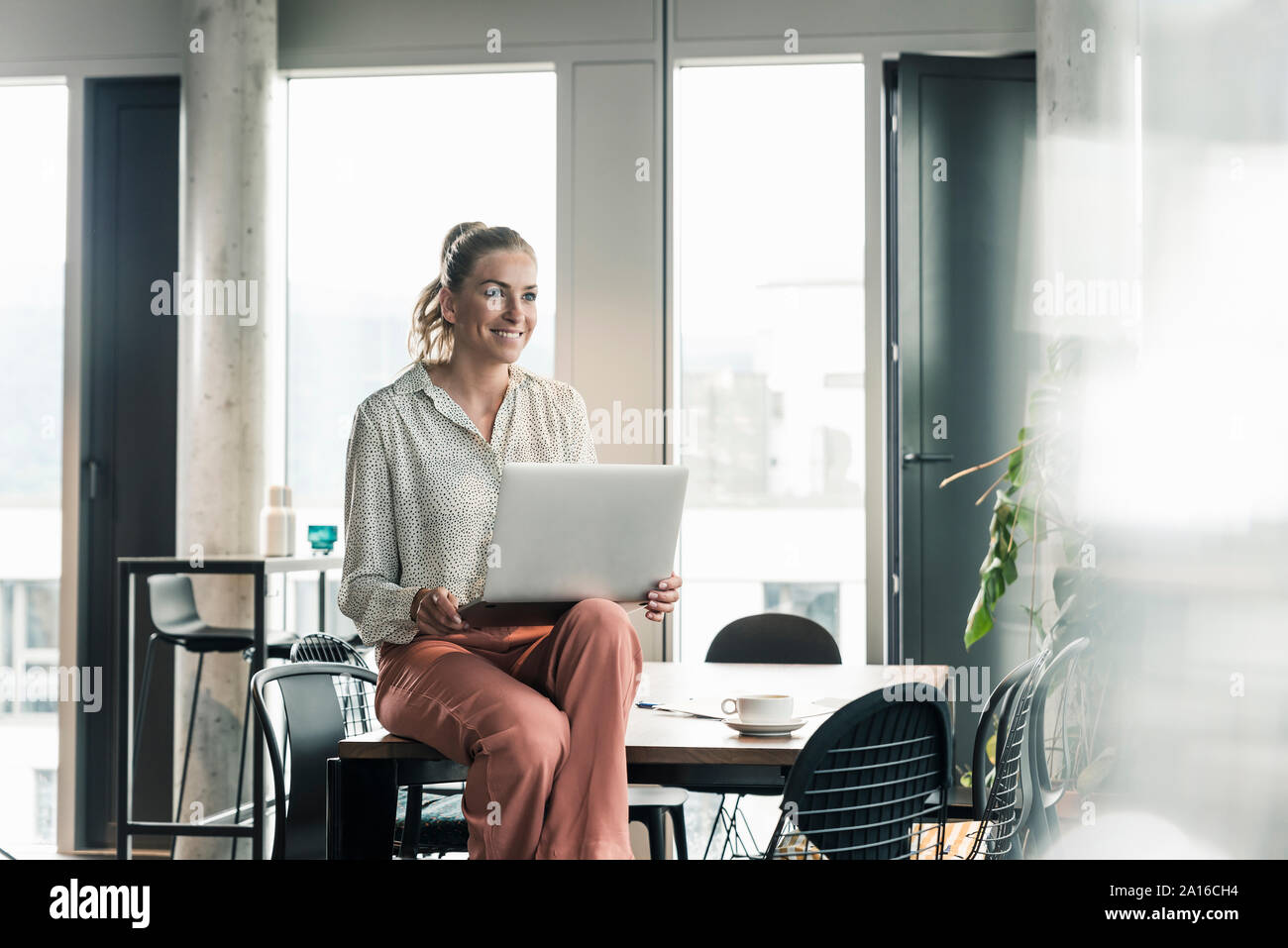 Sorridente imprenditrice seduta sul tavolo in ufficio con computer portatile Foto Stock