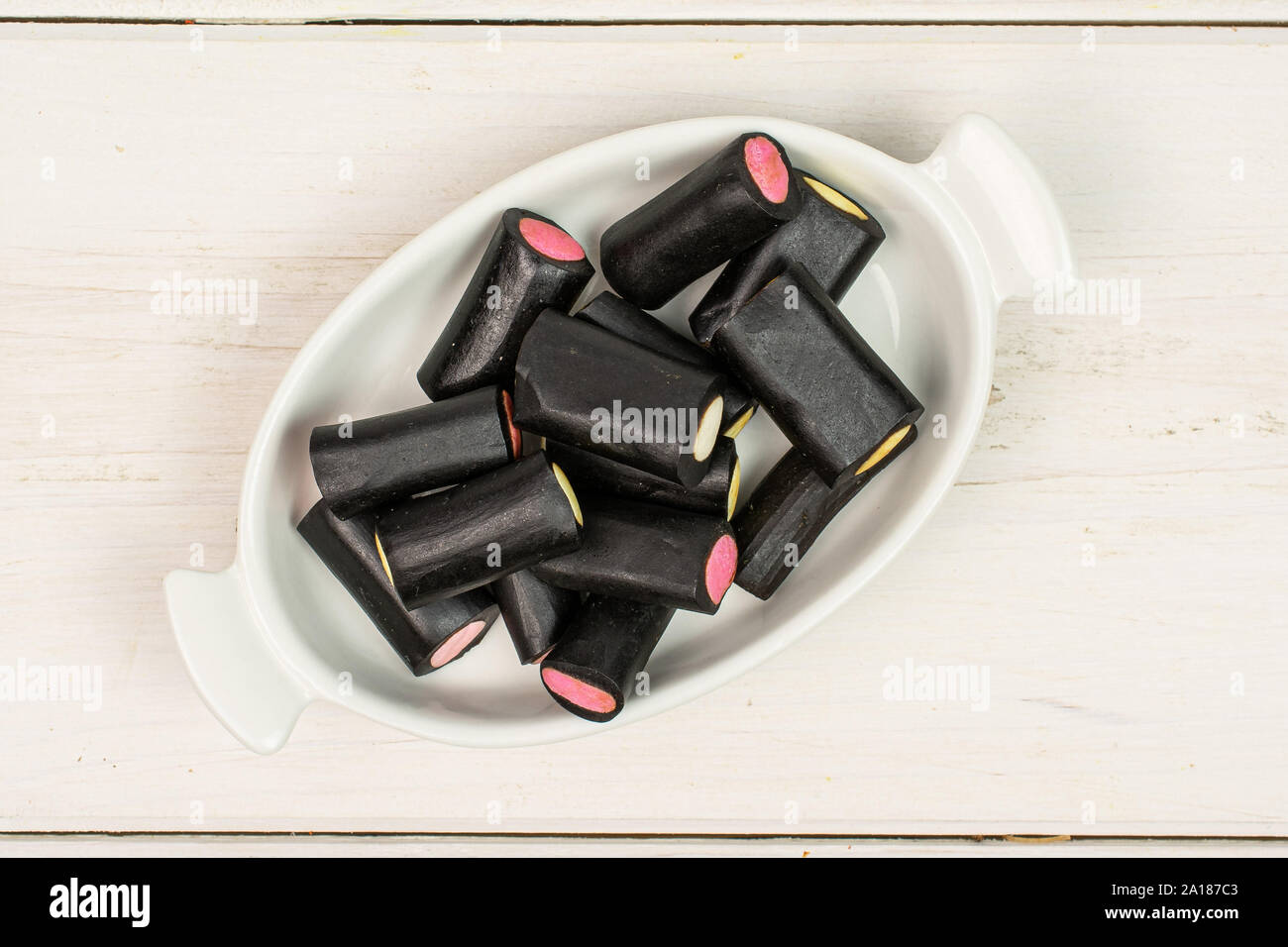 Sacco di tutto il dolce di liquirizia nera candy in bianco ovale tazza ceramica flatlay su legno bianco Foto Stock