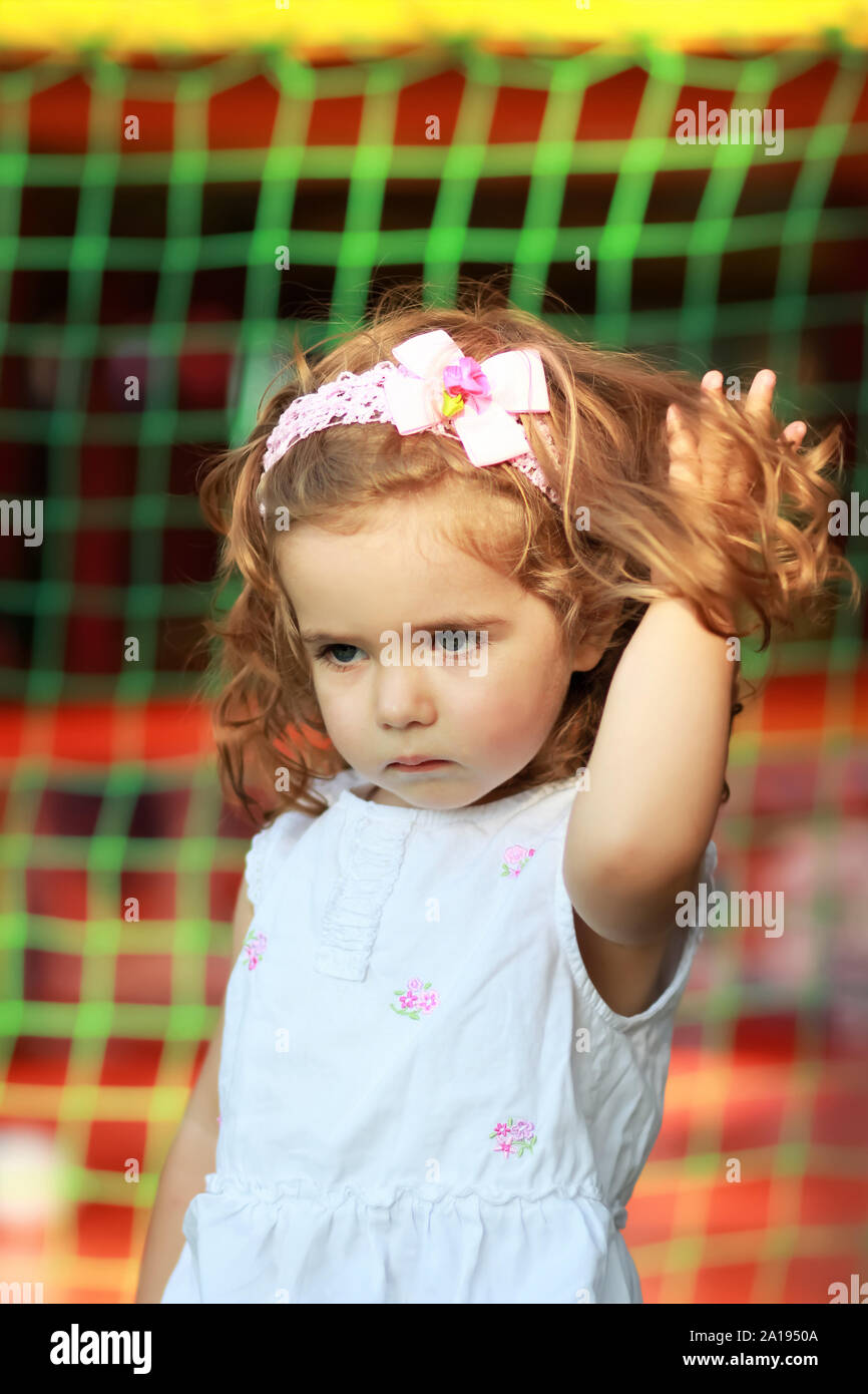 Ritratto di un dolce un anno vecchio ragazza con pink head band con archetto e fiore in estate park, mano bloccato in lunghi capelli ricci Foto Stock