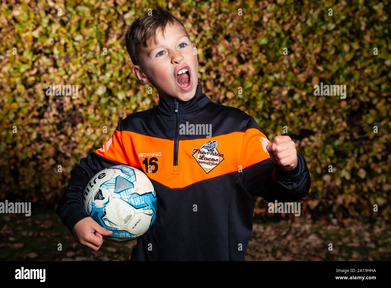 Un bambino di otto anni che indossa una striscia di calcio e tenendo un calcio sotto il suo braccio con l'altra mano in un pugno. Foto Stock