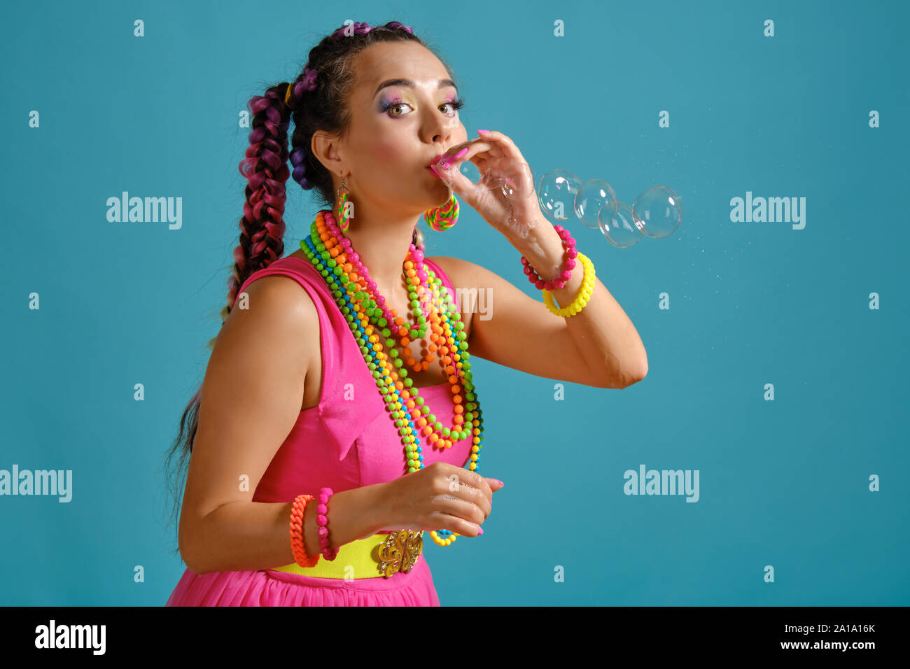 Bella ragazza con un multi-colore trecce acconciatura e luminoso per il make-up, è soffiare bolle usando le mani, in posa di studio contro uno sfondo blu. Foto Stock