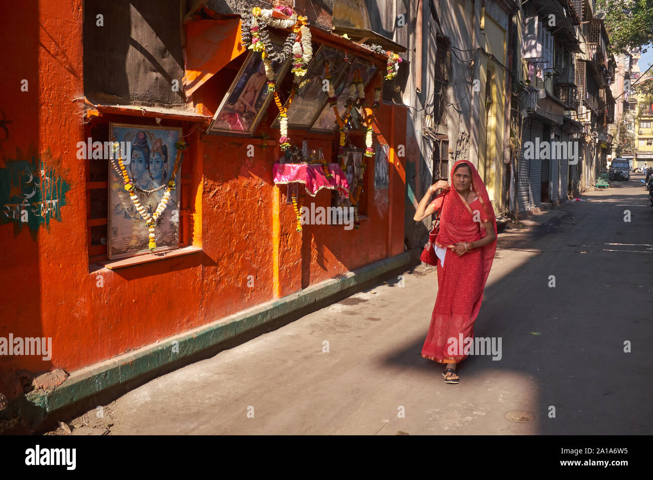 Una Donna vestita di un sari rosso passa una parete arancione e un indù santuario in una corsia in Mumbai, India; il colore arancione o 'saffron' è il colore dell'Induismo Foto Stock Una Donna vestita di un sari rosso passa una parete arancione e un indù santuario in una corsia in Mumbai, India; il colore arancione o 'saffron' è il colore dell'Induismo Foto Stock