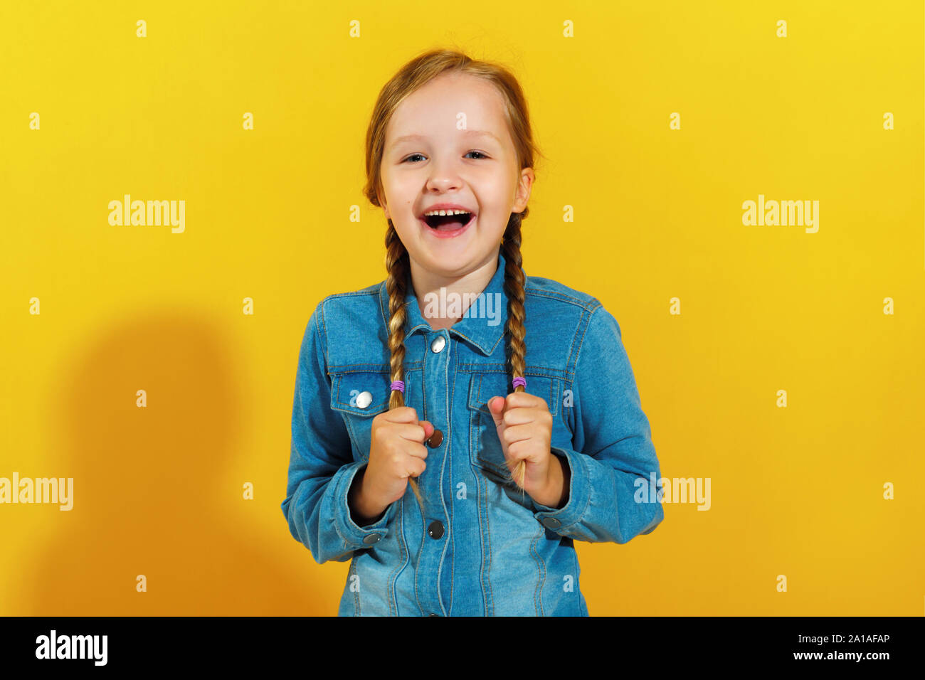 Allegro felice bambina in una camicia di denim su sfondo giallo. Il bambino ride e trattiene trecce di capelli. Foto Stock