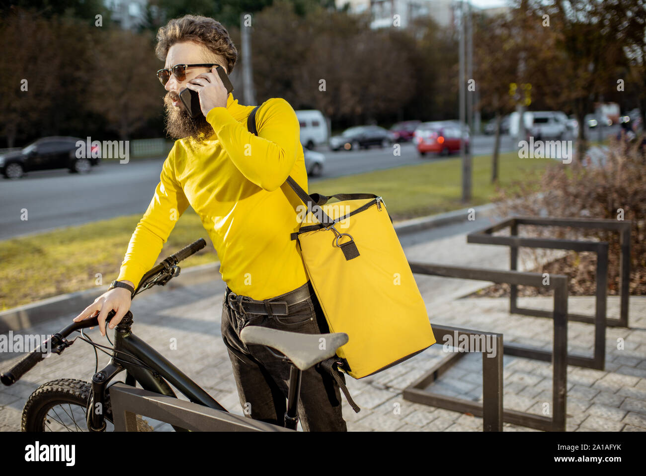 Giovani barbuto corriere a consegnare il cibo nella borsa termica su una bicicletta, parlando telefono con un client sulla strada di città Foto Stock
