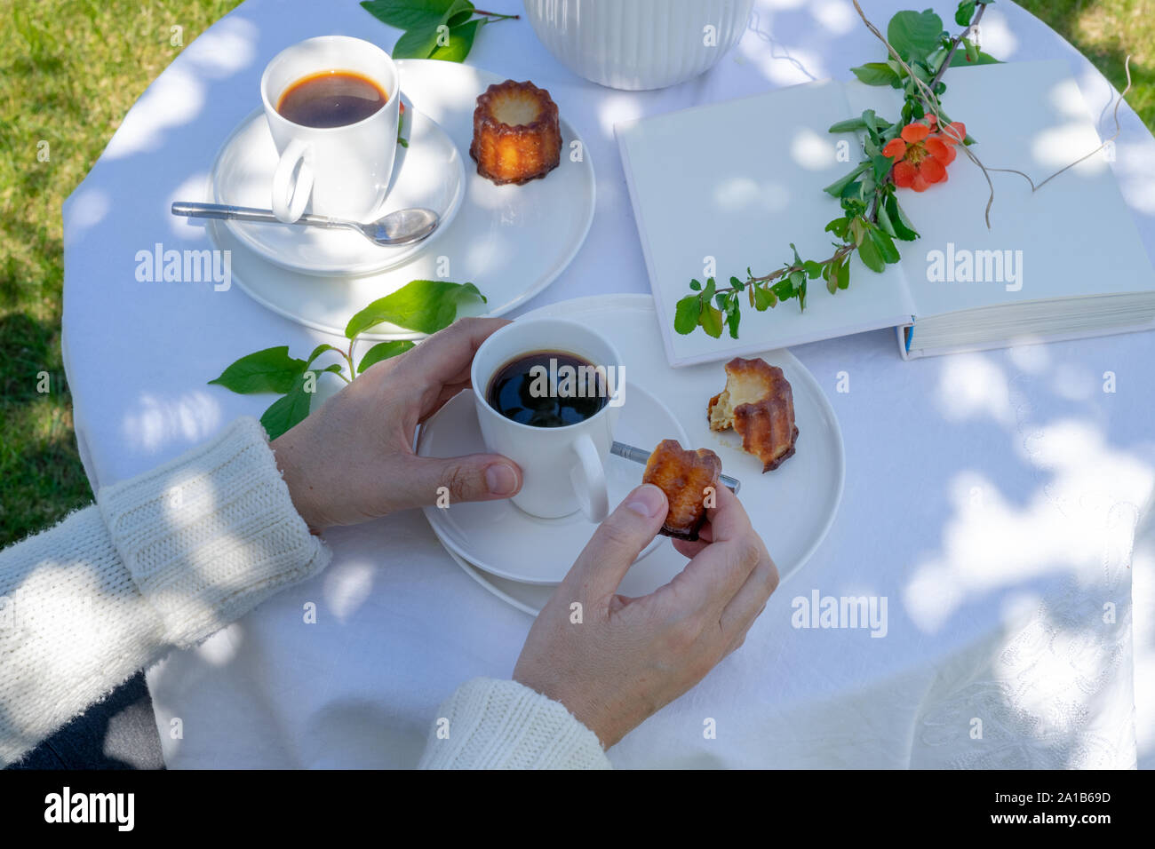 Bere un caffè di tempo in giardino. Servita tavolo con fiori, libro, caffè e dessert in giardino in primavera. Disposizione con una donna con le mani in mano. Foto Stock