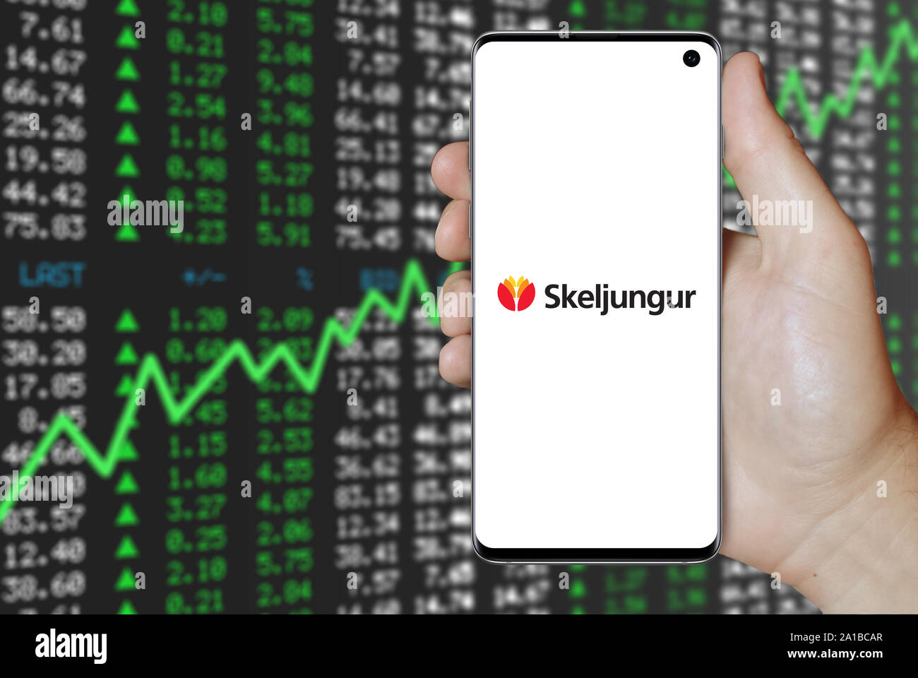Un uomo possiede uno smartphone visualizzando il logo della società Skeljungur quotata al Nasdaq Islanda. Positivo del mercato azionario sfondo. Credito: PIXDUCE Foto Stock