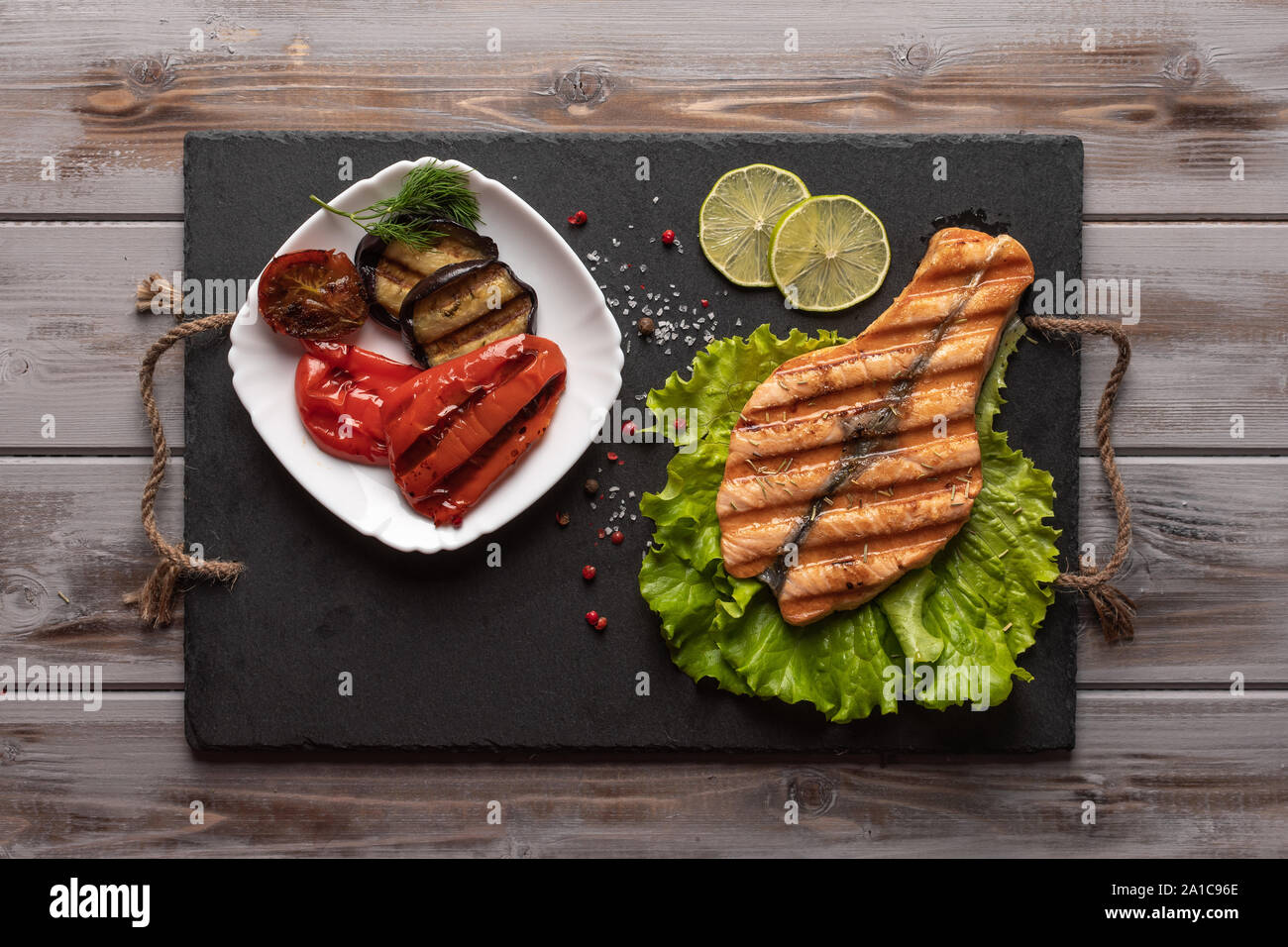 In casa ai ferri Bistecca di salmone su foglie di lattuga. Su un bordo scuro. Accanto alle verdure arrosto nella piastra, calce. Cibo sano. Vista superiore Foto Stock