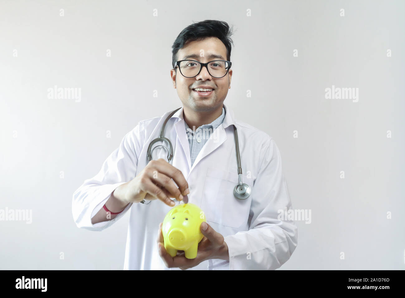 Indiano maschio medico in camice bianco e stetoscopio mettendo penny coin in un giallo salvadanaio Foto Stock