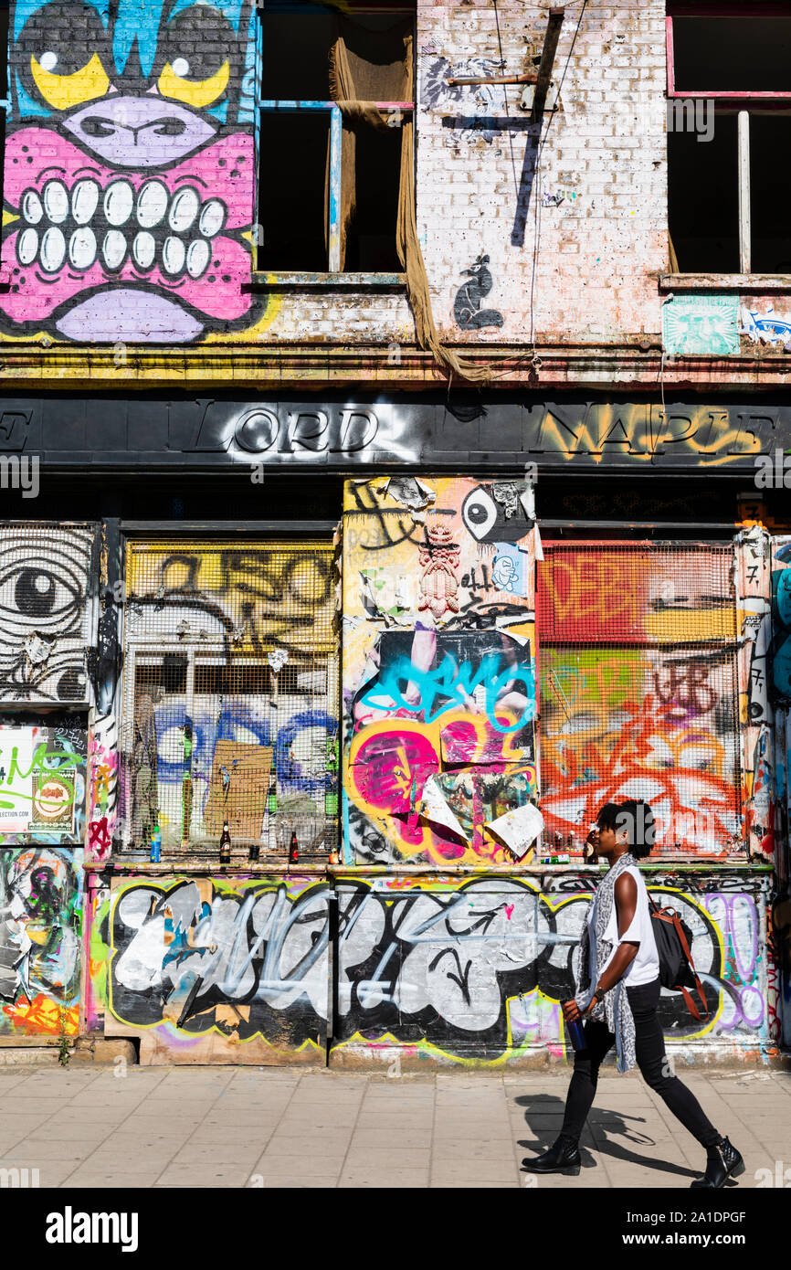 Una giovane donna cammina davanti a un edificio abbandonato coperto di graffiti in Hackney Wick, East London, Regno Unito. Foto Stock