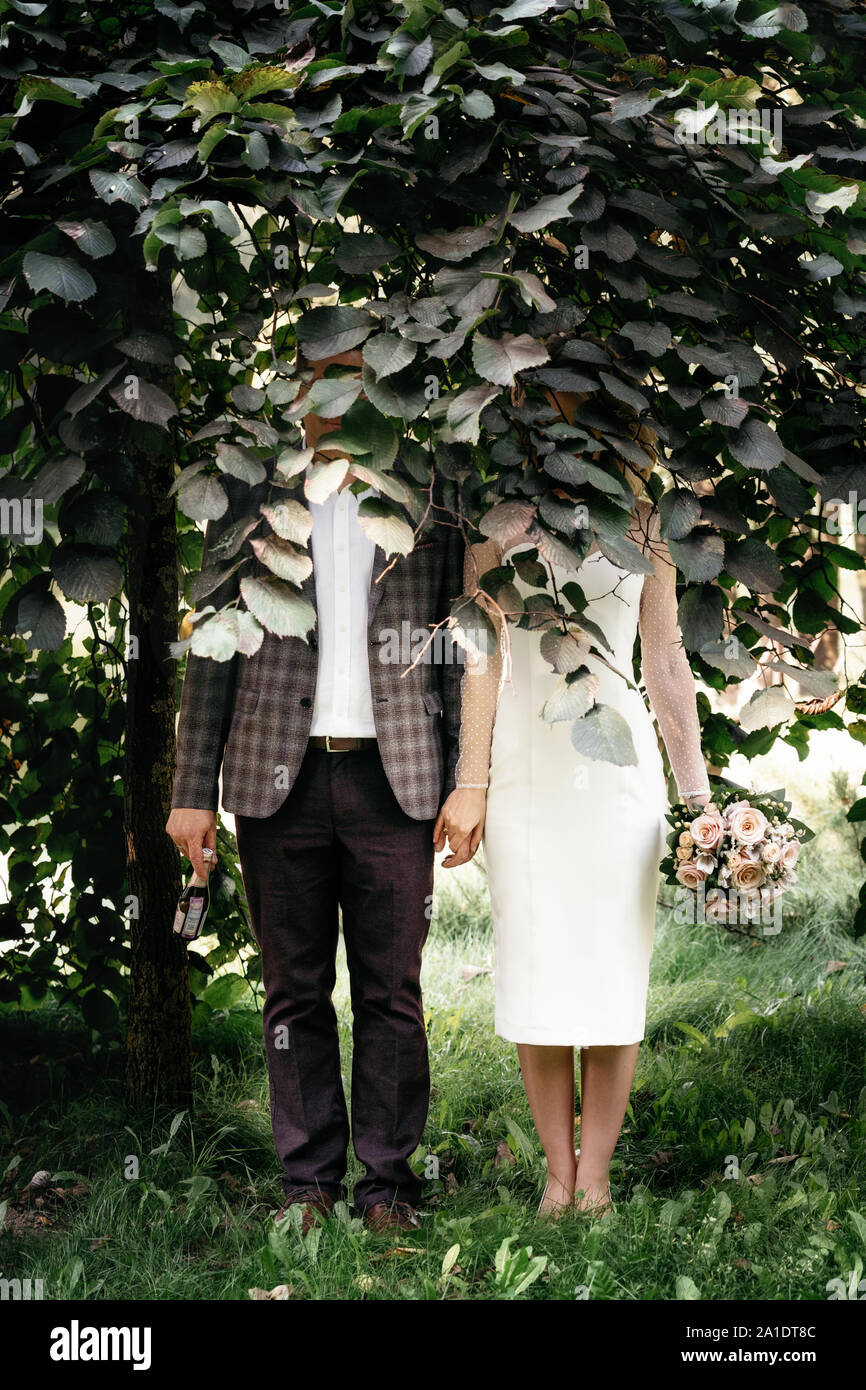 Coppie in viaggio di nozze dei cavalletti sotto albero verde con volti nascosti da fogliame e tenere le mani Foto Stock