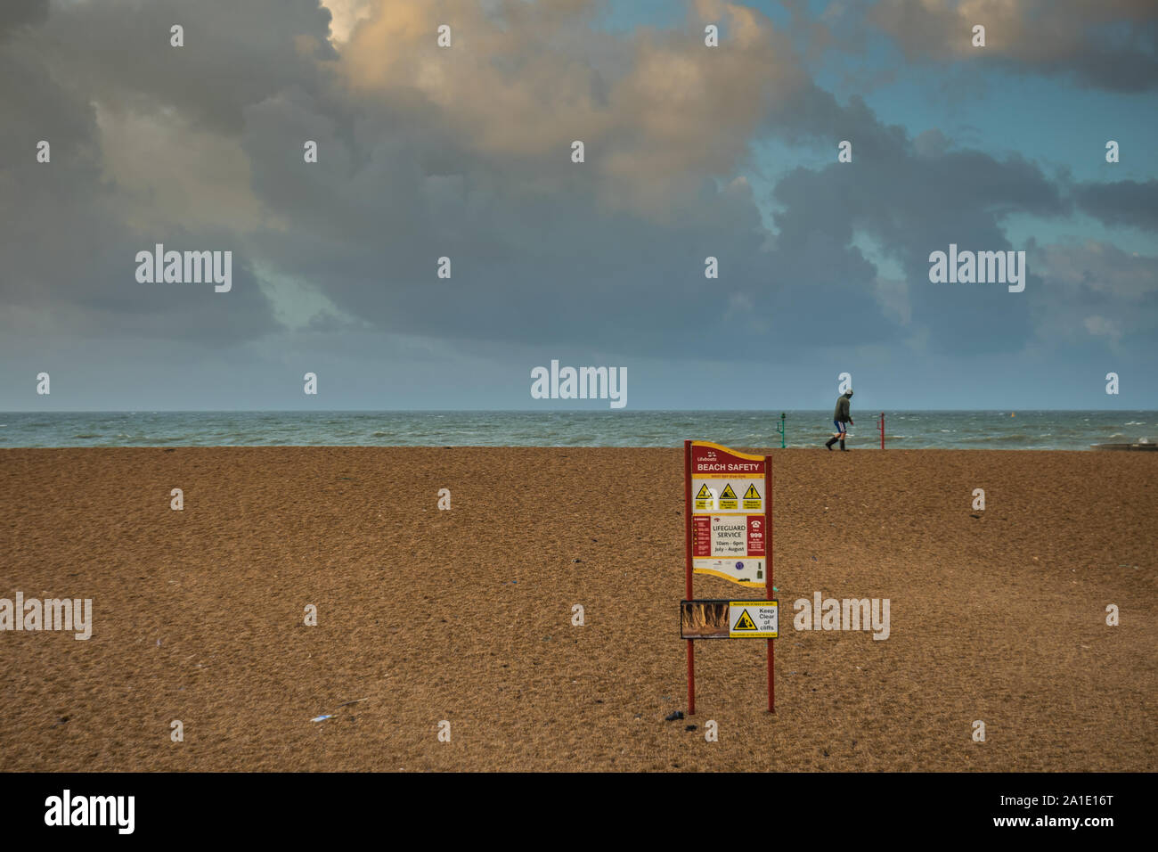 Persona con cappuccio passeggiate sulla spiaggia a ventoso; in primo piano un segno per la sicurezza in spiaggia Foto Stock
