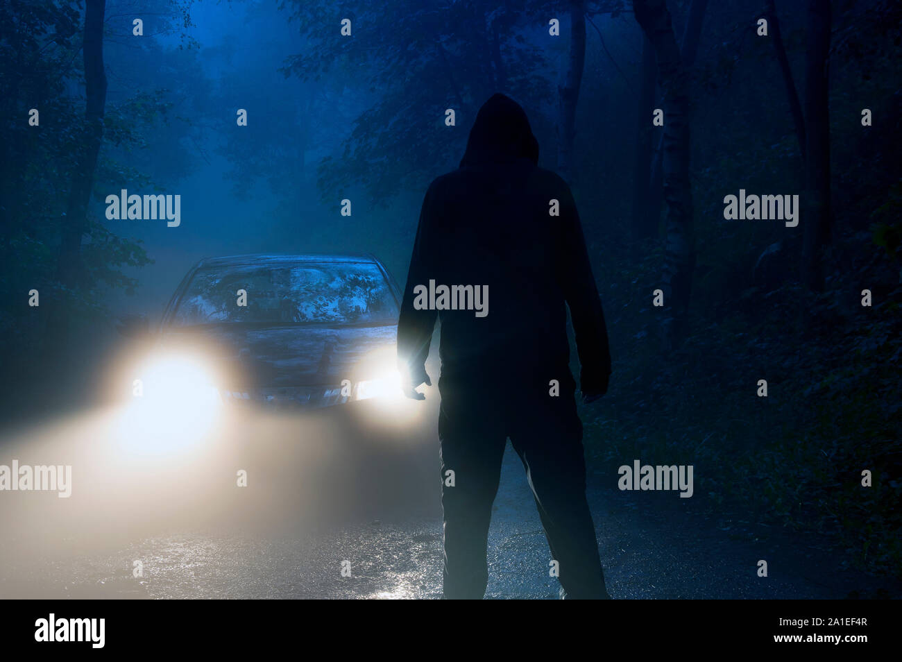 Un lone incappucciati figura in piedi nella parte anteriore dei fari delle auto nel mezzo di una strada di campagna su un losco, inquietanti, moody notte di nebbia Foto Stock