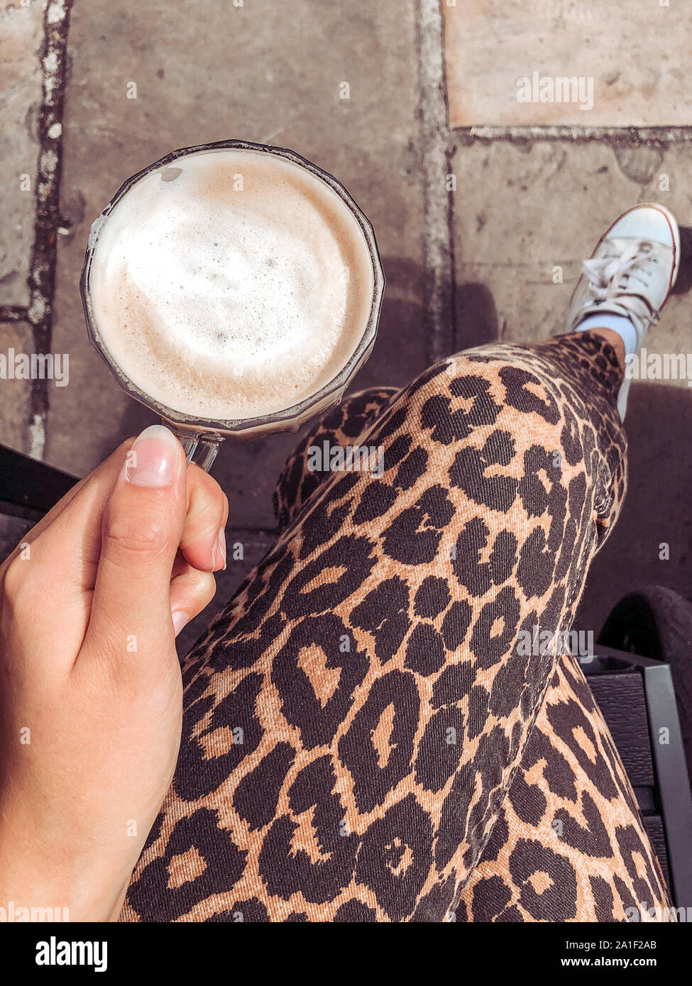 Donna seduta al di fuori cafe leopard indossa pantaloni di stampa tenendo un cappuccino al caffè da sopra Foto Stock