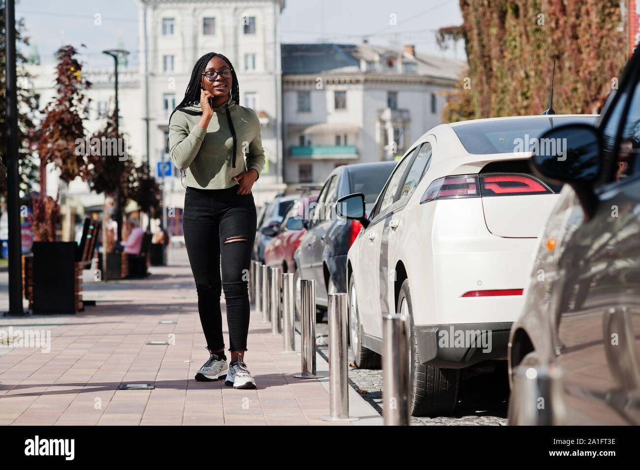 Città Ritratto di giovane positivo scuro femmina pelle indossando felpa con cappuccio verde e occhiali a piedi a parcheggio auto con telefono cellulare. Foto Stock