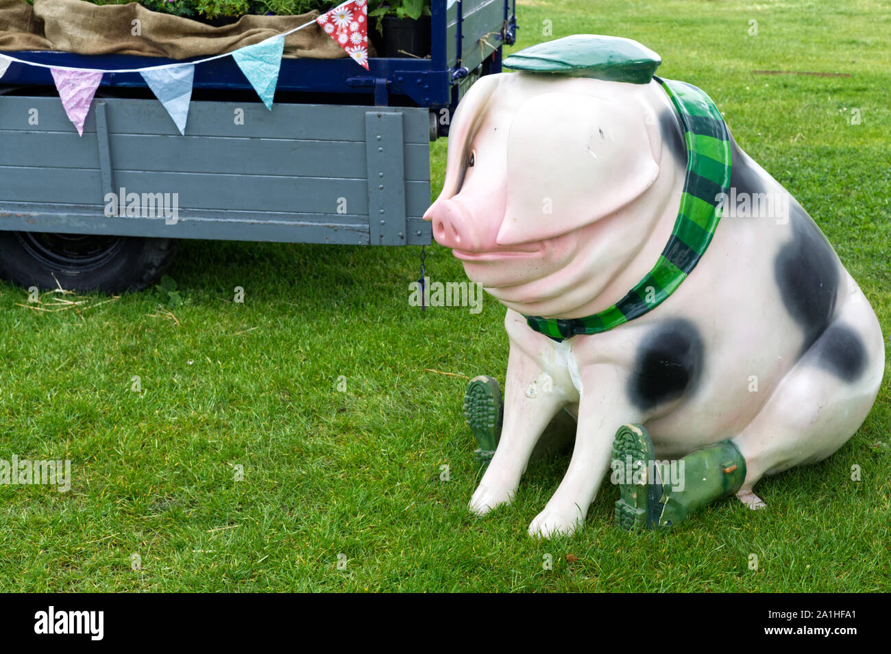 Statua di un suino a Wellington stivali, sciarpa e cappello seduto da un carrello con bunting Foto Stock